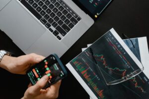 Manos sosteniendo un celular con datos financieros junto a una laptop con gráficas del mercado para comprar acciones en México desde una app