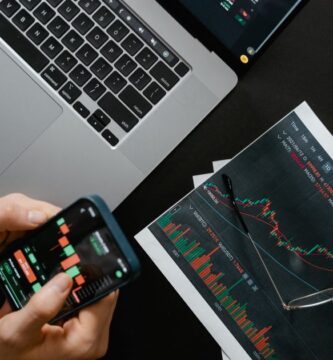 Manos sosteniendo un celular con datos financieros junto a una laptop con gráficas del mercado para comprar acciones en México desde una app