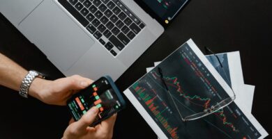 Manos sosteniendo un celular con datos financieros junto a una laptop con gráficas del mercado para comprar acciones en México desde una app