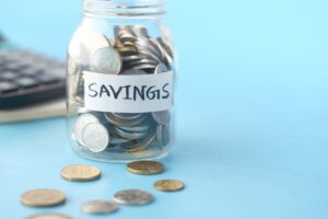 Frasco de vidrio etiquetado como ahorro lleno de monedas junto a una calculadora, para calcular cuánto necesitas en tu fondo de emergencia