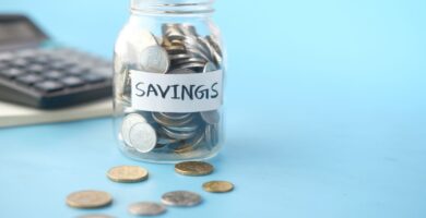Frasco de vidrio etiquetado como ahorro lleno de monedas junto a una calculadora, para calcular cuánto necesitas en tu fondo de emergencia