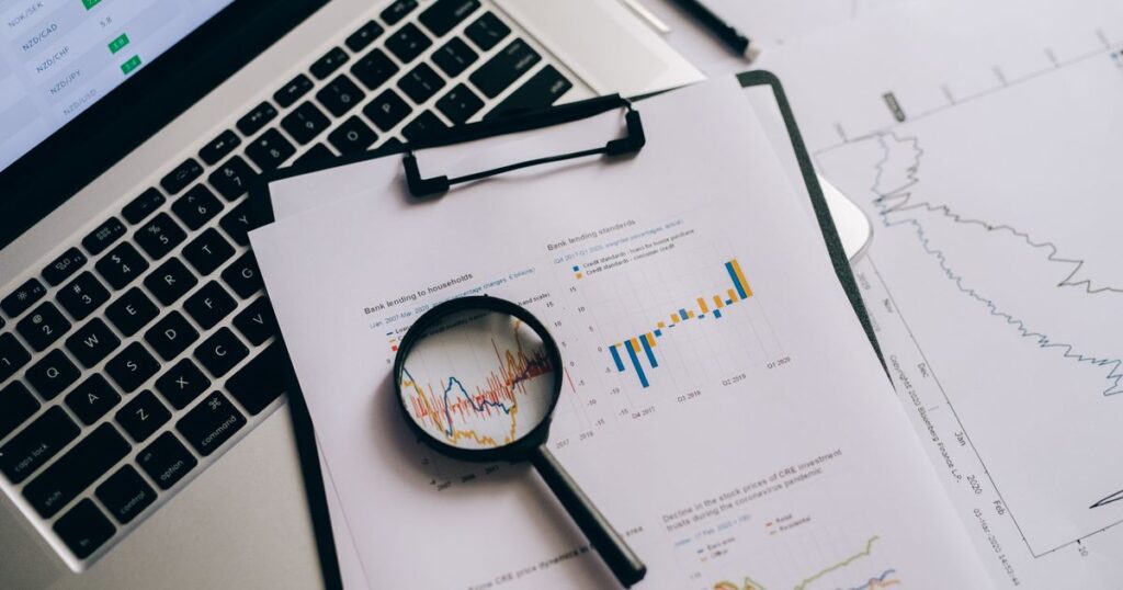 Lupa sobre gráficos financieros impresos junto a una laptop con cotizaciones, analizando los datos en busca de un fraude de inversión