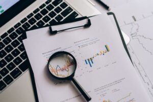 Lupa sobre gráficos financieros impresos junto a una laptop con cotizaciones, analizando los datos en busca de un fraude de inversión