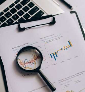 Lupa sobre gráficos financieros impresos junto a una laptop con cotizaciones, analizando los datos en busca de un fraude de inversión
