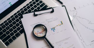 Lupa sobre gráficos financieros impresos junto a una laptop con cotizaciones, analizando los datos en busca de un fraude de inversión