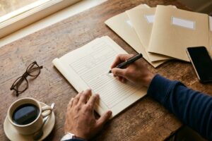 Manos de una persona llenando a mano un formato de designación de beneficiarios con una pluma fuente, sobre un escritorio de madera junto a la ventana, con tres carpetas de archivo cerradas y etiquetadas, unos lentes, una taza de café y un celular boca abajo