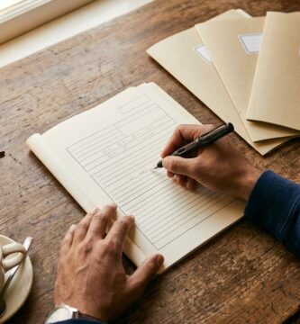 Manos de una persona llenando a mano un formato de designación de beneficiarios con una pluma fuente, sobre un escritorio de madera junto a la ventana, con tres carpetas de archivo cerradas y etiquetadas, unos lentes, una taza de café y un celular boca abajo