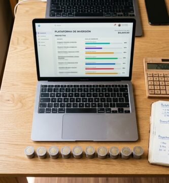 Mesa de madera vista desde arriba con una laptop que muestra un panel de proyectos de inversión, una fila de doce montoncitos pequeños de monedas mexicanas, una calculadora y una libreta con anotaciones