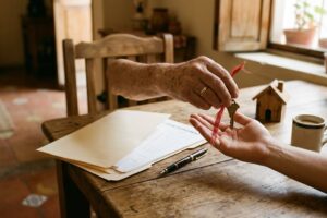 Mano de una persona mayor con argolla de matrimonio entregando una llave de casa atada con un listón rojo en la palma abierta de una persona joven, sobre una mesa de madera con una carpeta de documentos, una pluma, una casita de madera y una taza de café