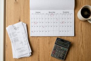 Vista cenital de un escritorio de madera con un calendario con fechas marcadas, una pila de recibos sujetos con un clip, una calculadora y una taza de café