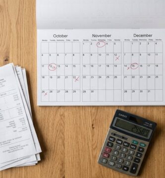Vista cenital de un escritorio de madera con un calendario con fechas marcadas, una pila de recibos sujetos con un clip, una calculadora y una taza de café