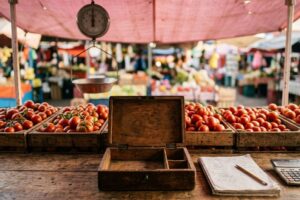 Mostrador de madera de un puesto de jitomates en un mercado mexicano con una caja de dinero de madera abierta y vacía, una libreta con apuntes a lápiz y una calculadora, con huacales de jitomate y una báscula colgante al fondo